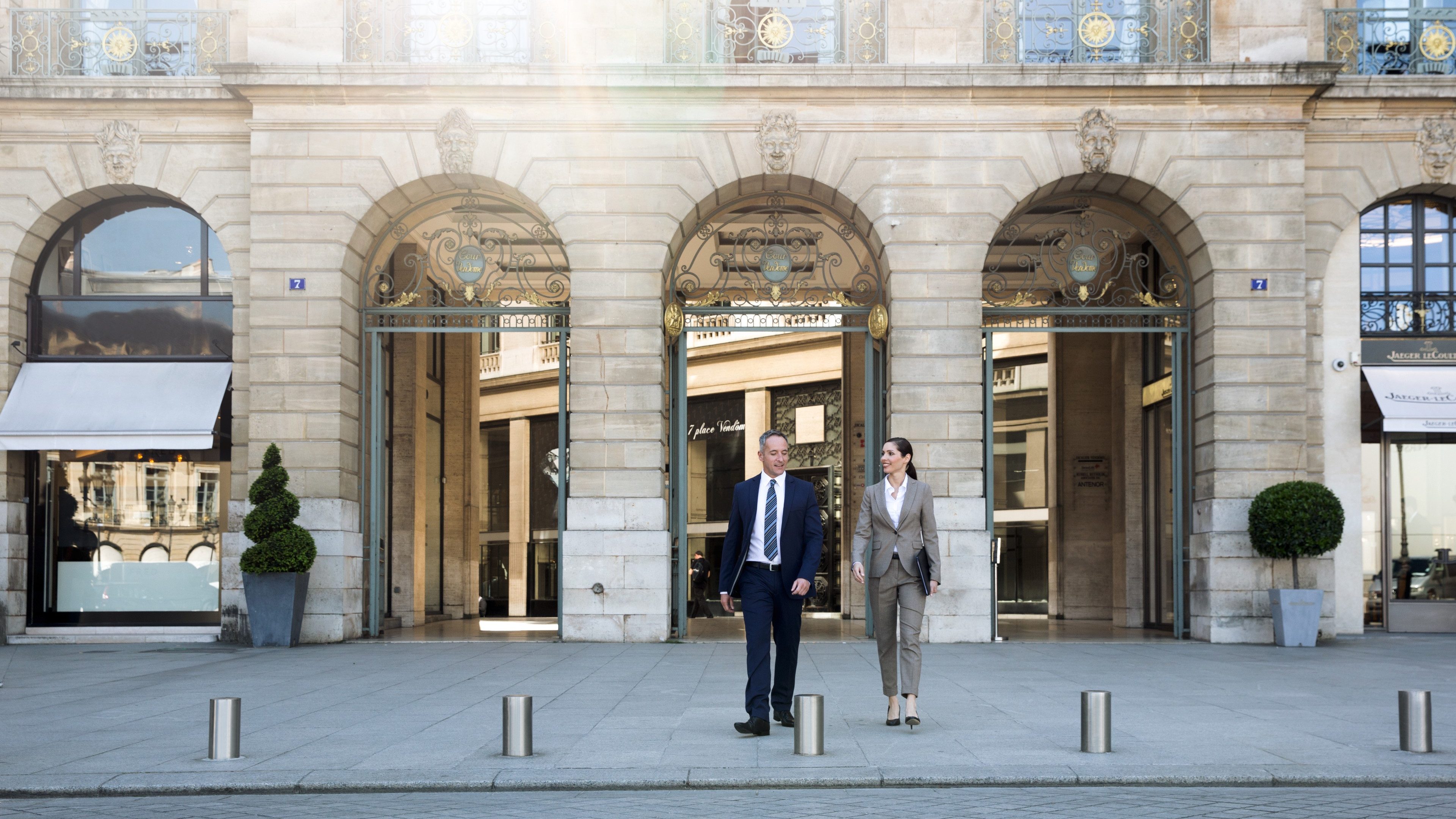 Mann und Frau in Businesskleidung vor einem repräsentativen Gebäude, in dessen Fenstern die Sonne reflektiert. 