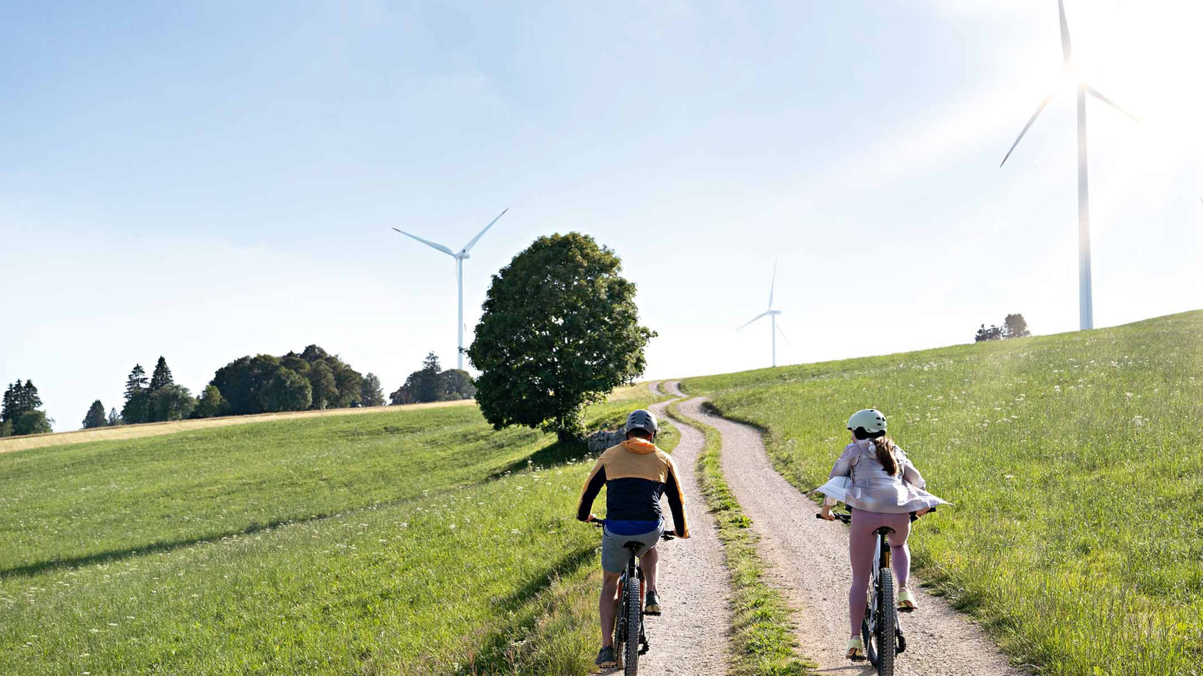 Zwei Personen fahren mit dem Fahrrad auf einer unbefestigten Straße, im Hintergrund sind Windräder zu sehen.