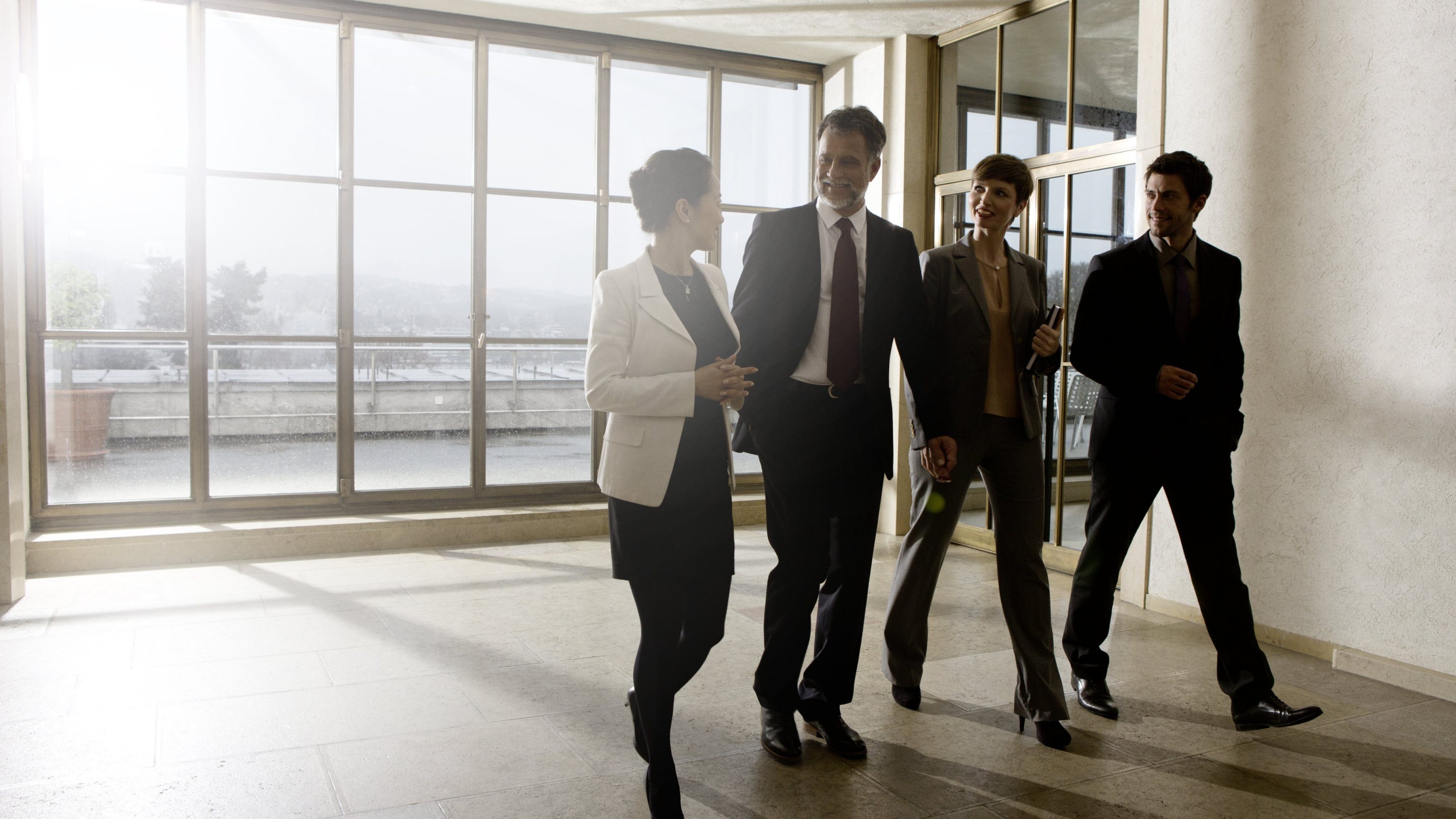 Two women and two men in business attire, standing in front of a floor-to-ceiling lattice window and looking out through it. The sun shines into the room.
