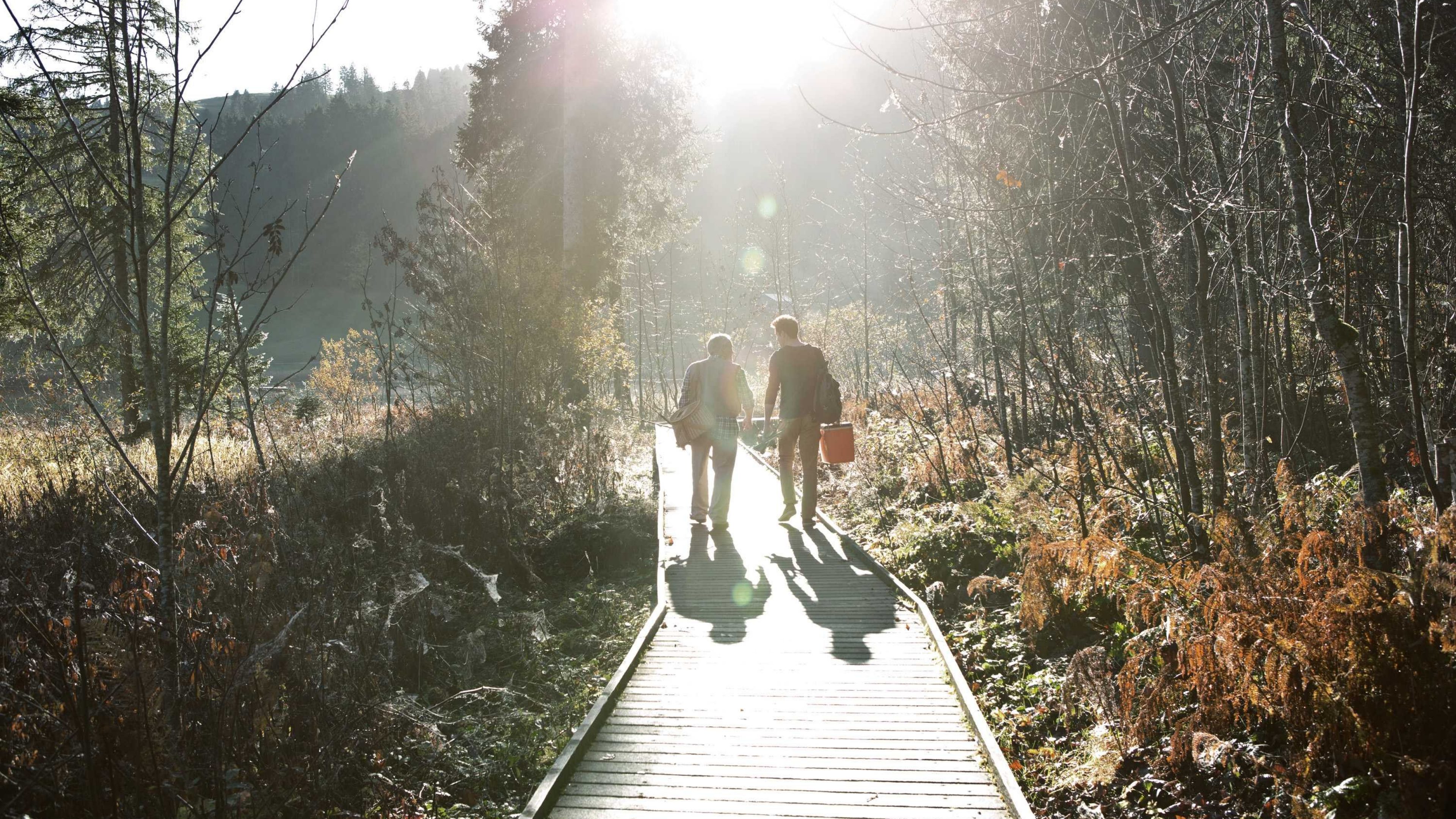 Two people walking along a wooden path in nature, both carrying fishing equipment in their hands.