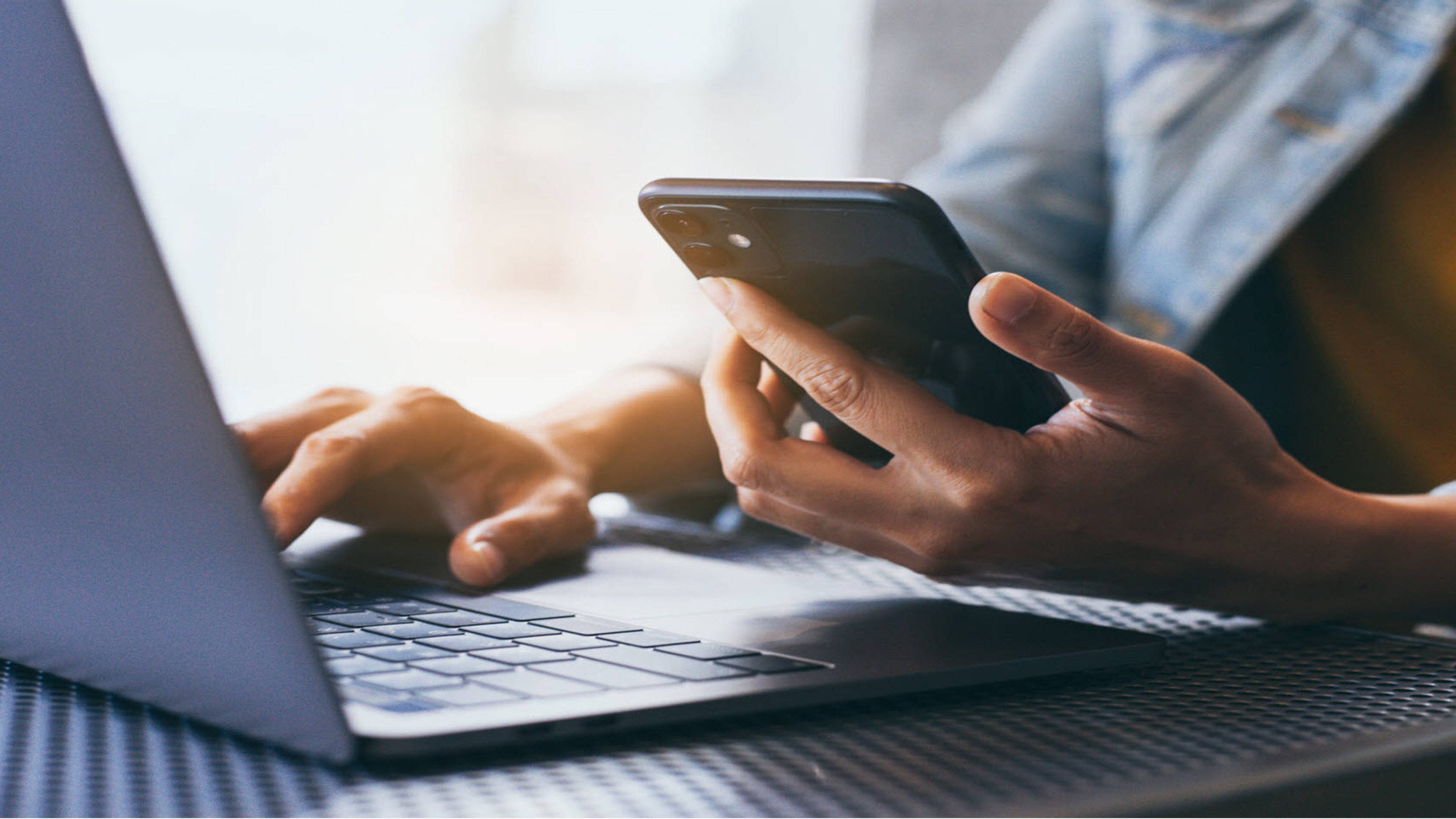 A person sitting at a laptop with a mobile phone in their hand.