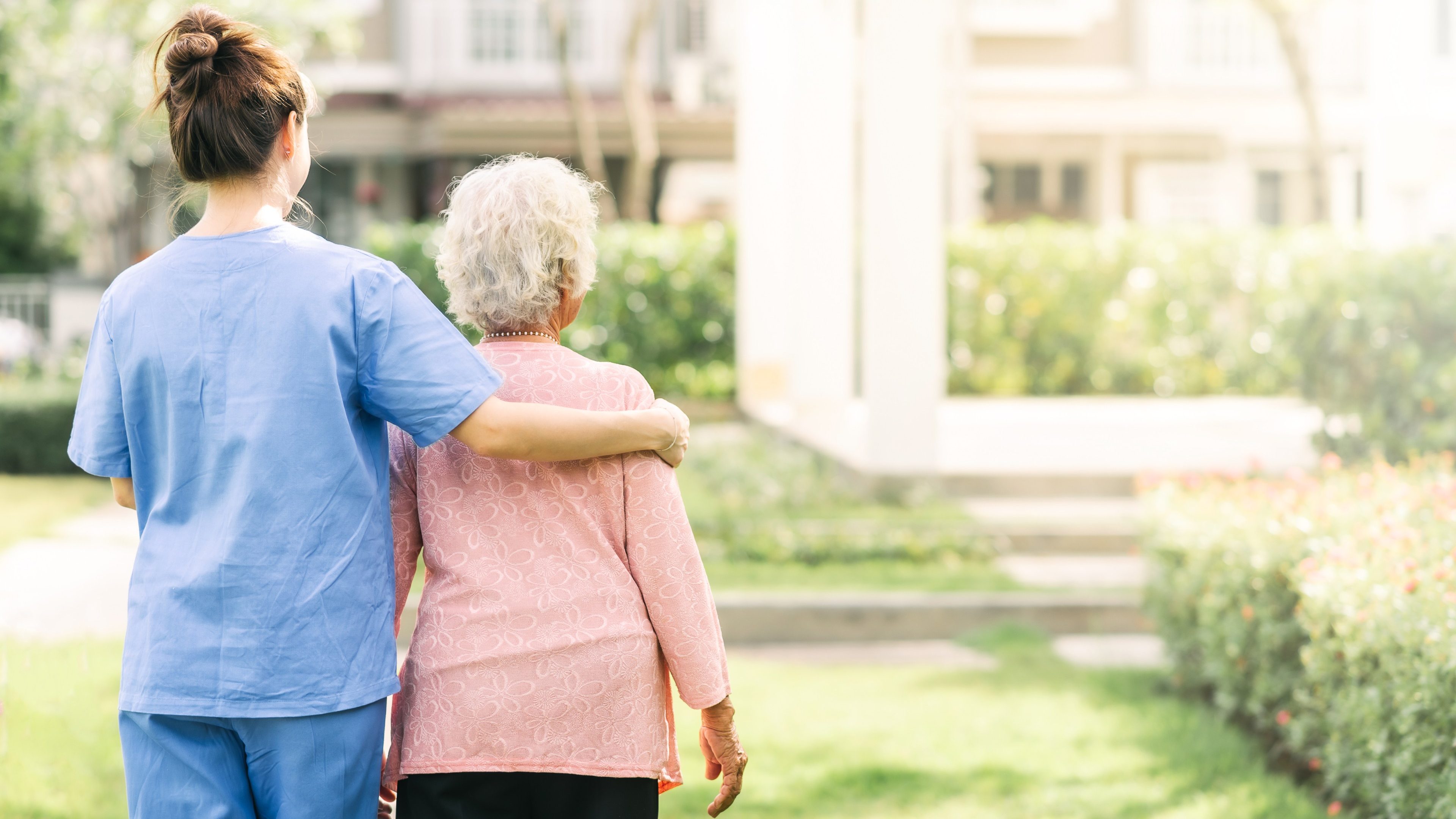Une infirmière met son bras autour d’une dame âgée. Toutes deux observent un jardin.