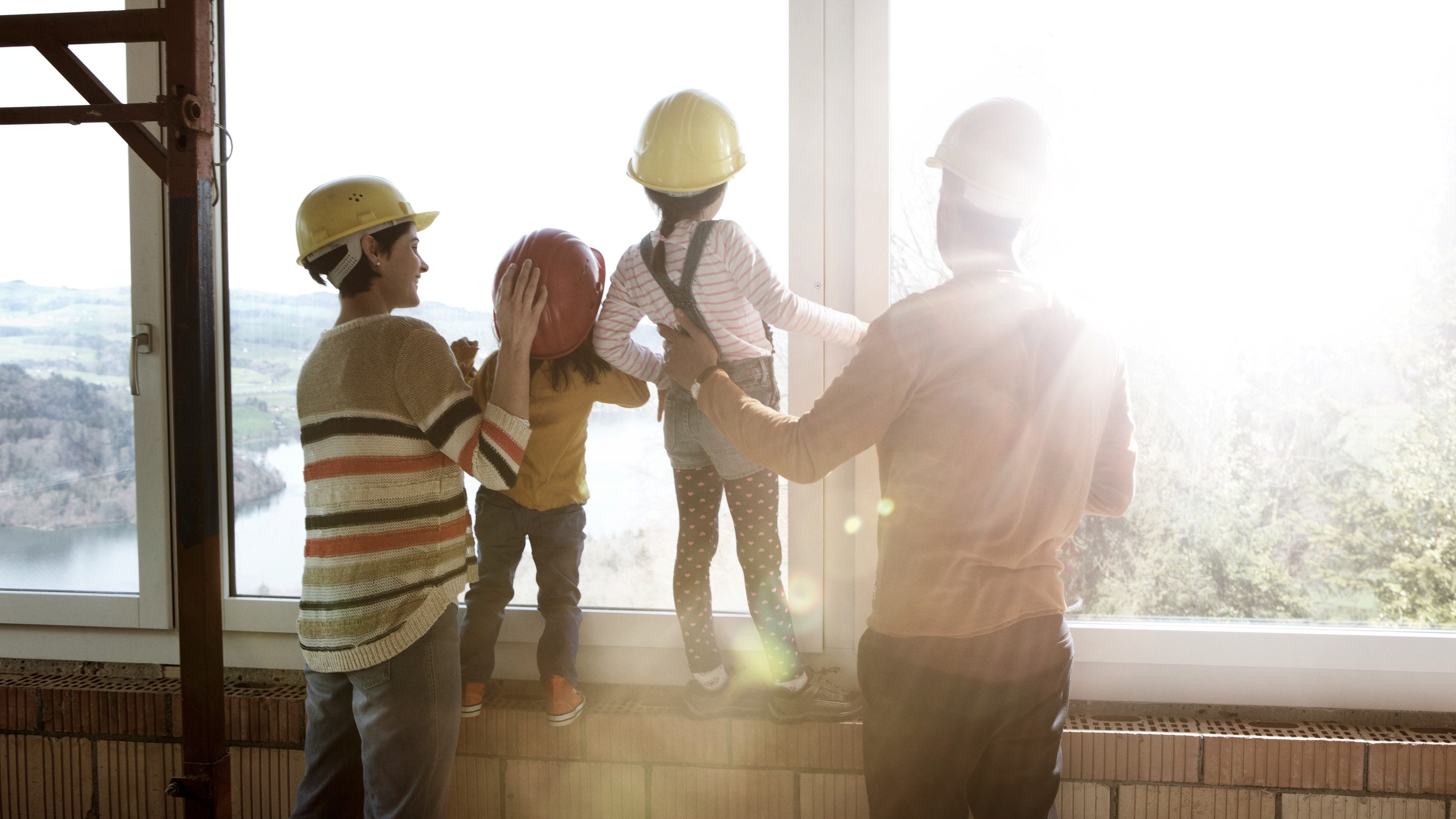 Jeune famille de quatre personnes avec un casque de protection, debout sur une embrasure et regardant par la fenêtre. Le soleil rayonne vers eux et vers le lecteur.